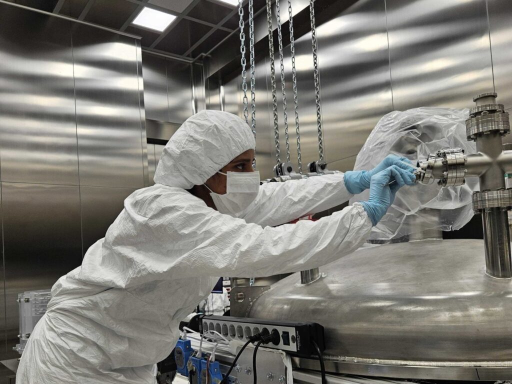 A researcher in cleanroom protective clothing adjusting a large metallic scientific apparatus inside a laboratory environment.