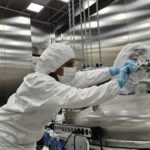 A researcher in cleanroom protective clothing adjusting a large metallic scientific apparatus inside a laboratory environment.