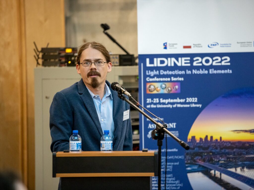 Scientist speaking at a podium during an international physics conference, with a microphone, water bottles, and a “LIDINE 2022 – Light Detection in Noble Elements” conference banner in the background.