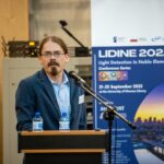Scientist speaking at a podium during an international physics conference, with a microphone, water bottles, and a “LIDINE 2022 – Light Detection in Noble Elements” conference banner in the background.