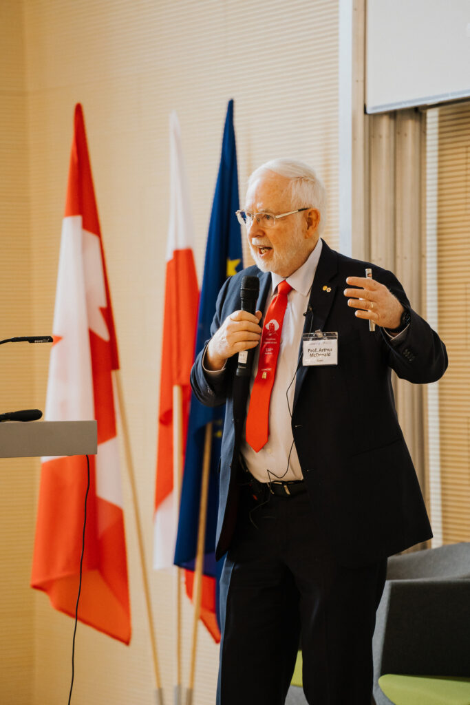 A man in a suit and red tie speaking into a handheld microphone during a formal event, gesturing with one hand, with national and European Union flags visible in the background.