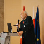 Scientist speaking at a podium during an international physics conference, with a microphone and a conference banner visible in the background.