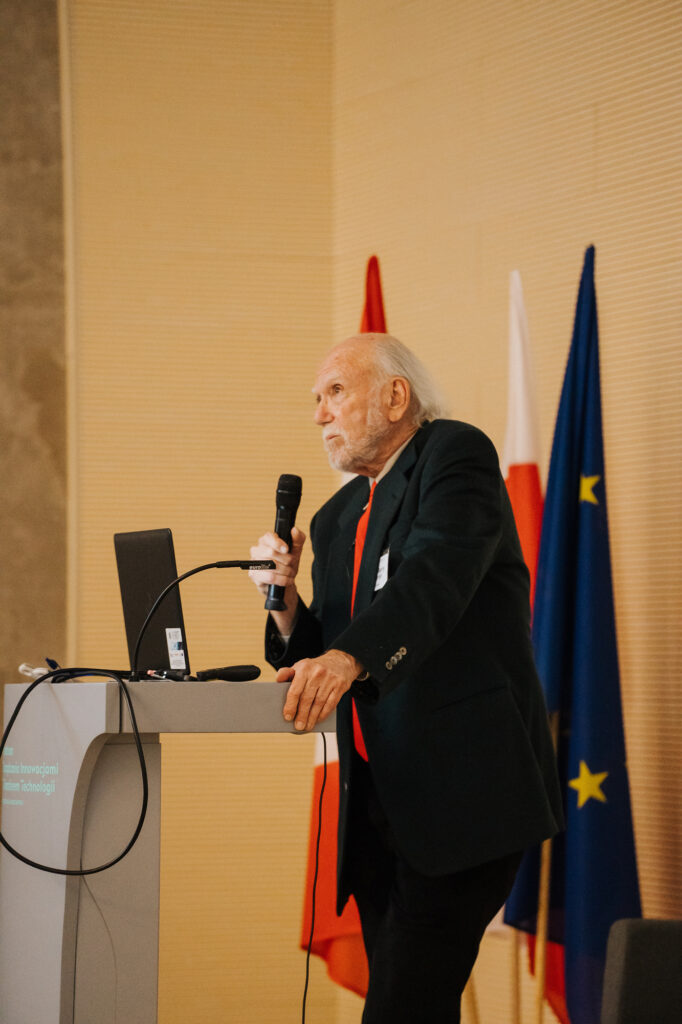 Scientist speaking at a podium during an international physics conference, with a microphone and a conference banner visible in the background.
