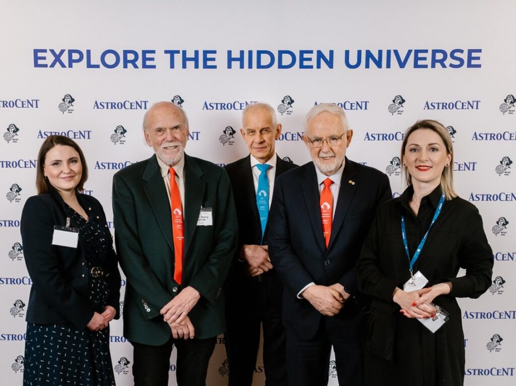 Five people pose for a photo in front of an AstroCeNT-branded backdrop with the slogan “EXPLORE THE HIDDEN UNIVERSE,” wearing conference badges and colored lanyards.