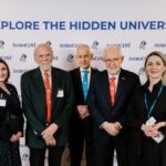 Five people pose for a photo in front of an AstroCeNT-branded backdrop with the slogan “EXPLORE THE HIDDEN UNIVERSE,” wearing conference badges and colored lanyards.