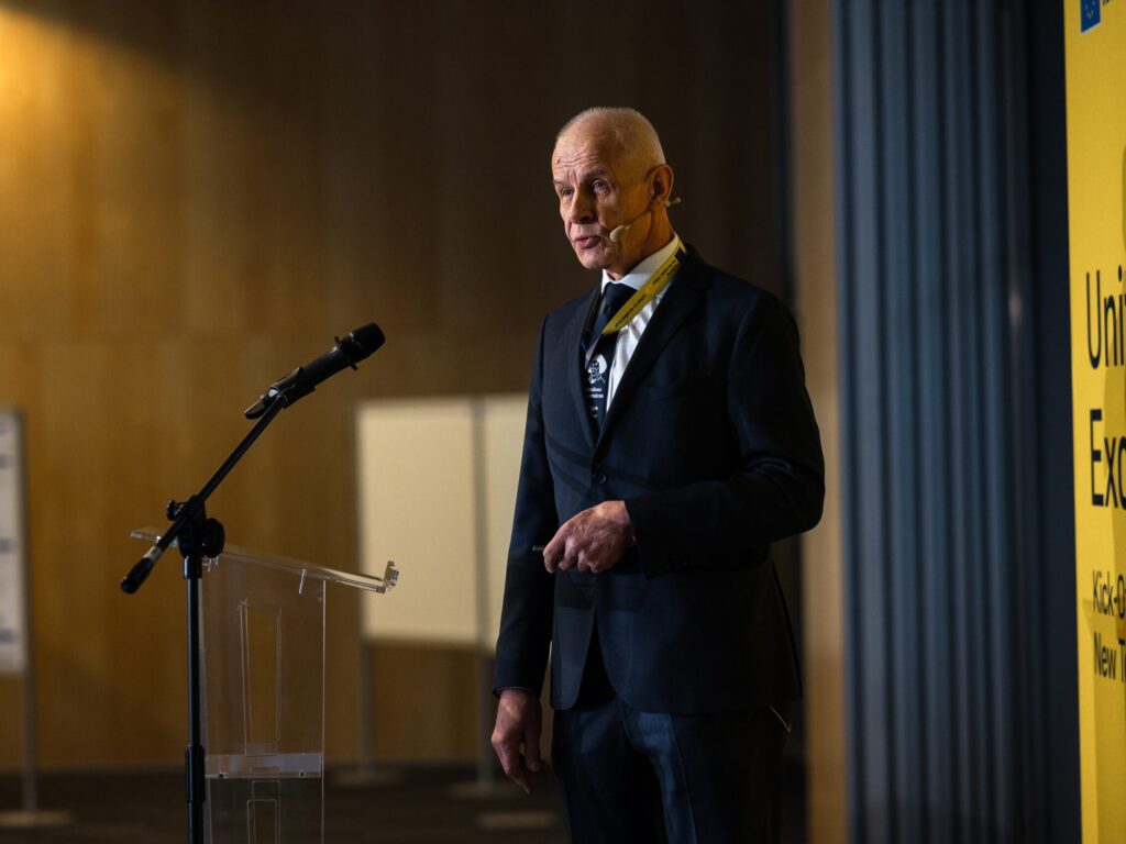A man in a dark suit speaks at a conference podium, wearing a headset microphone, with a yellow event banner partially visible to the right.