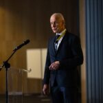 A man in a dark suit speaks at a conference podium, wearing a headset microphone, with a yellow event banner partially visible to the right.