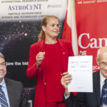 At a formal event, a man in a suit holds up two signed documents while a woman in a red suit stands behind him; an “AstroCeNT” banner and a large “Canada” backdrop are visible.