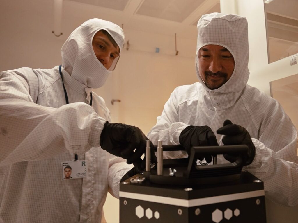 Two researchers in white cleanroom suits and black gloves work together to assemble or adjust a black scientific instrument inside a cleanroom.