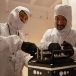 Two researchers in white cleanroom suits and black gloves work together to assemble or adjust a black scientific instrument inside a cleanroom.
