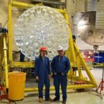 Two researchers in blue coveralls and hard hats stand in an industrial lab in front of a large circular array of clear detector modules mounted on a yellow support frame; a large metallic sphere and corrugated ducting are visible behind them.