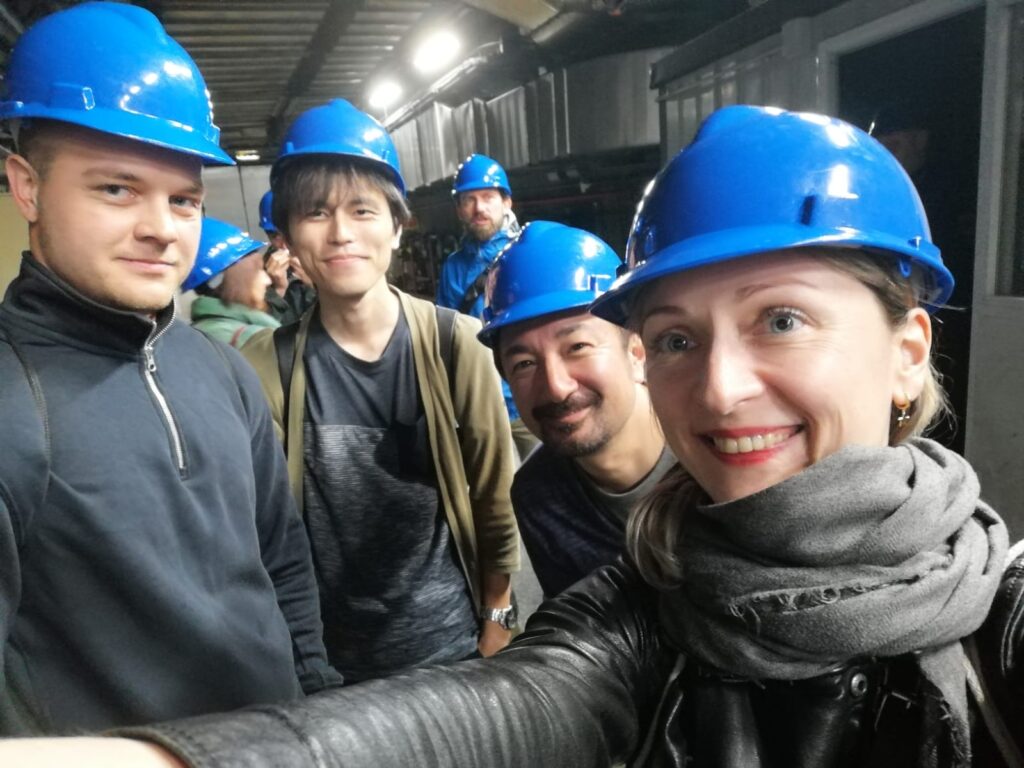 Group selfie of several people wearing blue hard hats inside an industrial underground facility or tunnel corridor.